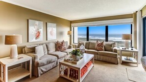 Una sala de estar con un ventanal que ofrece vistas al océano, un sofá beige, una mesa de centro y dos mesas auxiliares con lámparas.
