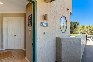A building entrance with a number sign '712', a door, a wall-mounted light, and a doormat. - 712-sailmaker (Fernandina Beach)