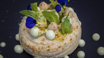 A dessert with a creamy texture, garnished with blue flowers, mint leaves, and small round balls, served on a dark plate.