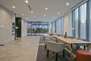Un espacio de oficina moderno con una larga mesa, sillas y un sofá. Hay ventanas amplias con cortinas, vistas a zonas verdes y una puerta con panel de vidrio.