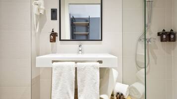 Un baño moderno con un lavamanos blanco, dos toallas blancas, un box de ducha de vidrio y dos botellas de champú.