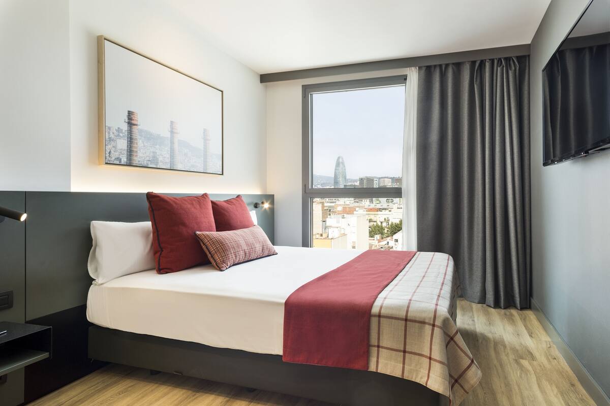Une chambre d’hôtel moderne avec un grand lit, une vue sur la ville, un téléviseur fixé au mur et un tableau encadré accroché au mur.