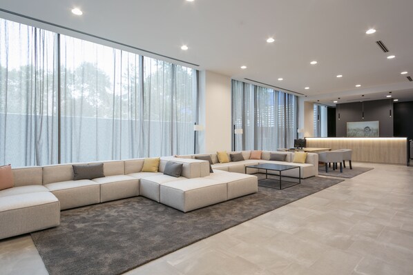 A spacious living area with a large sectional sofa, a coffee table, and a dining table. There are large windows with curtains, a kitchen area with a countertop, and a modern ceiling with recessed lighting.