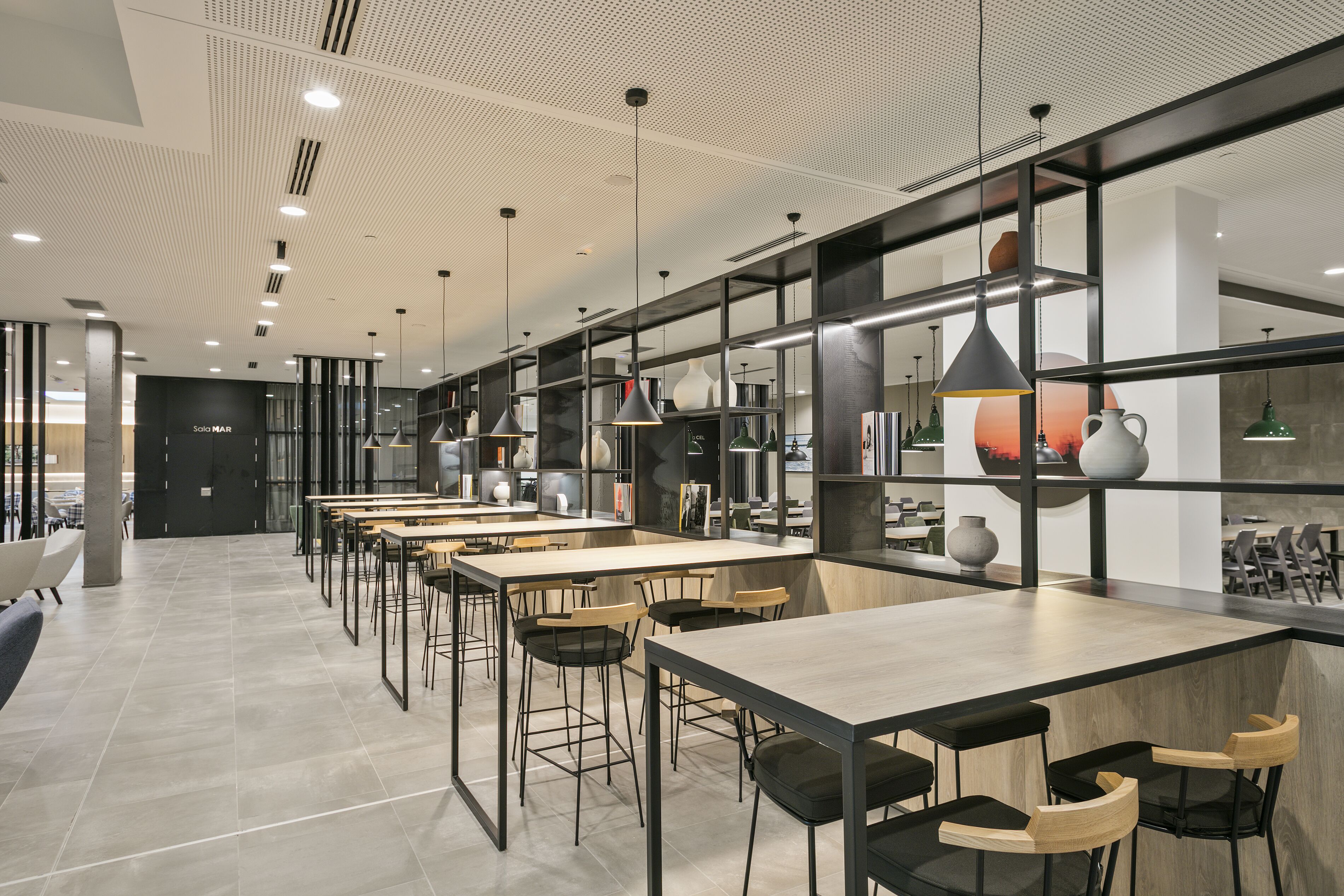 Zona pranzo moderna con tavoli e sedie lunghe, soffitto alto con illuminazione a incasso e un'ampia finestra che offre luce naturale.