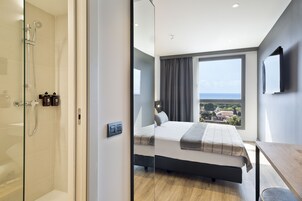Quarto de hotel moderno com uma cama grande, chuveiro em vidro, vista para a cidade e TV de tela plana na parede.