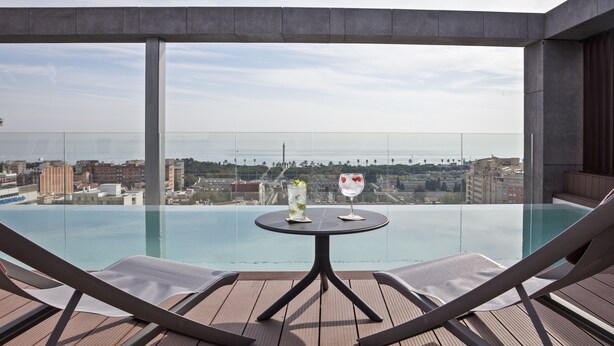 Una piscina infinita con una mesa y dos sillas reclinables, con vistas a un paisaje urbano.