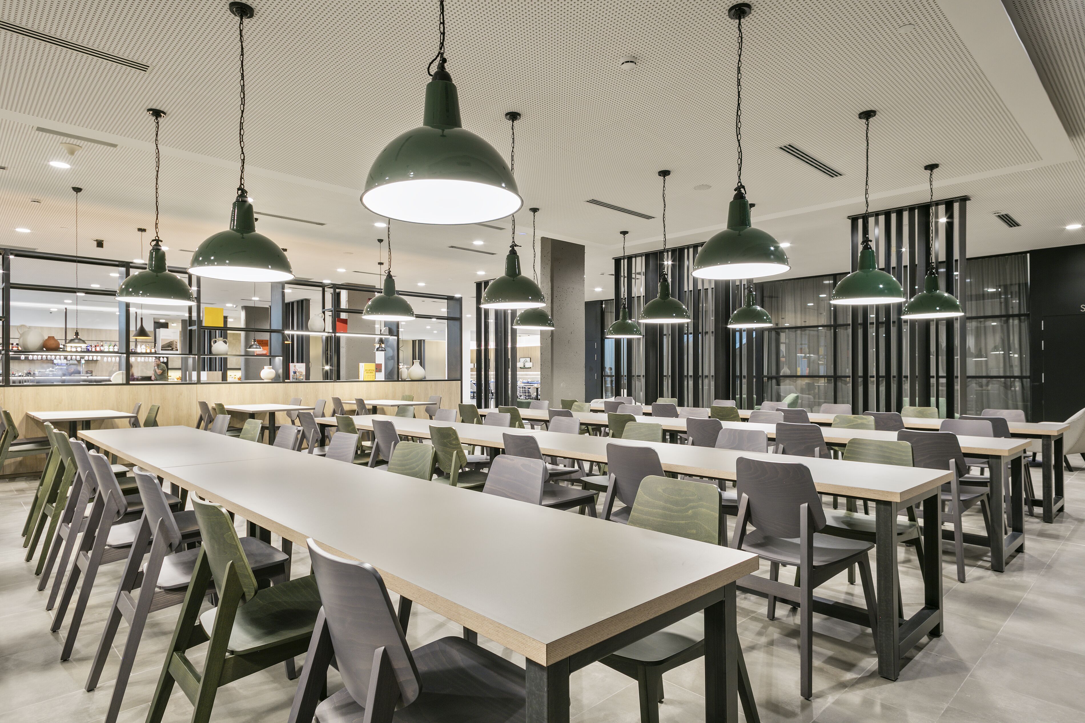 A modern dining area with long tables and chairs, green pendant lights, and a view of the kitchen area in the background.