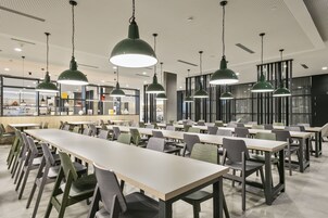 A modern dining area with long tables and chairs, green pendant lights, and a view of the kitchen area in the background.