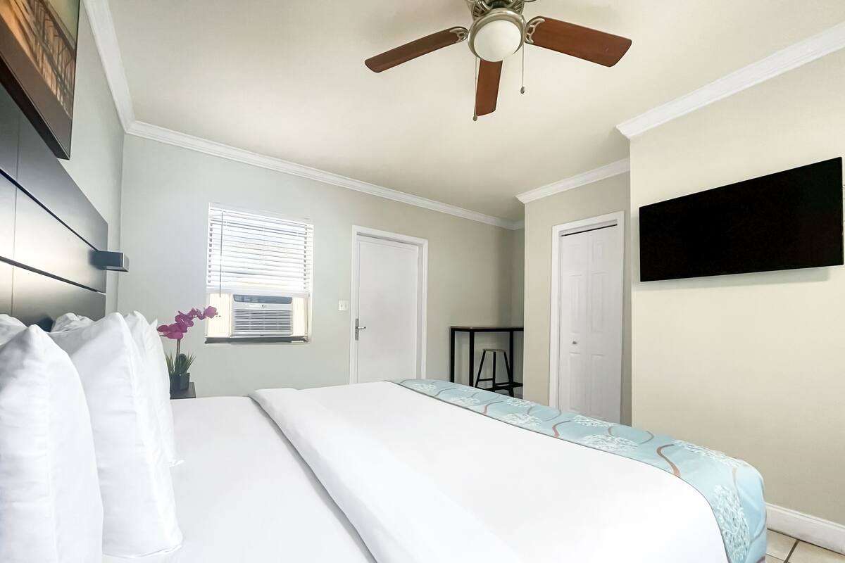 A bedroom with a bed, a ceiling fan, a window with blinds, and a TV mounted on the wall.