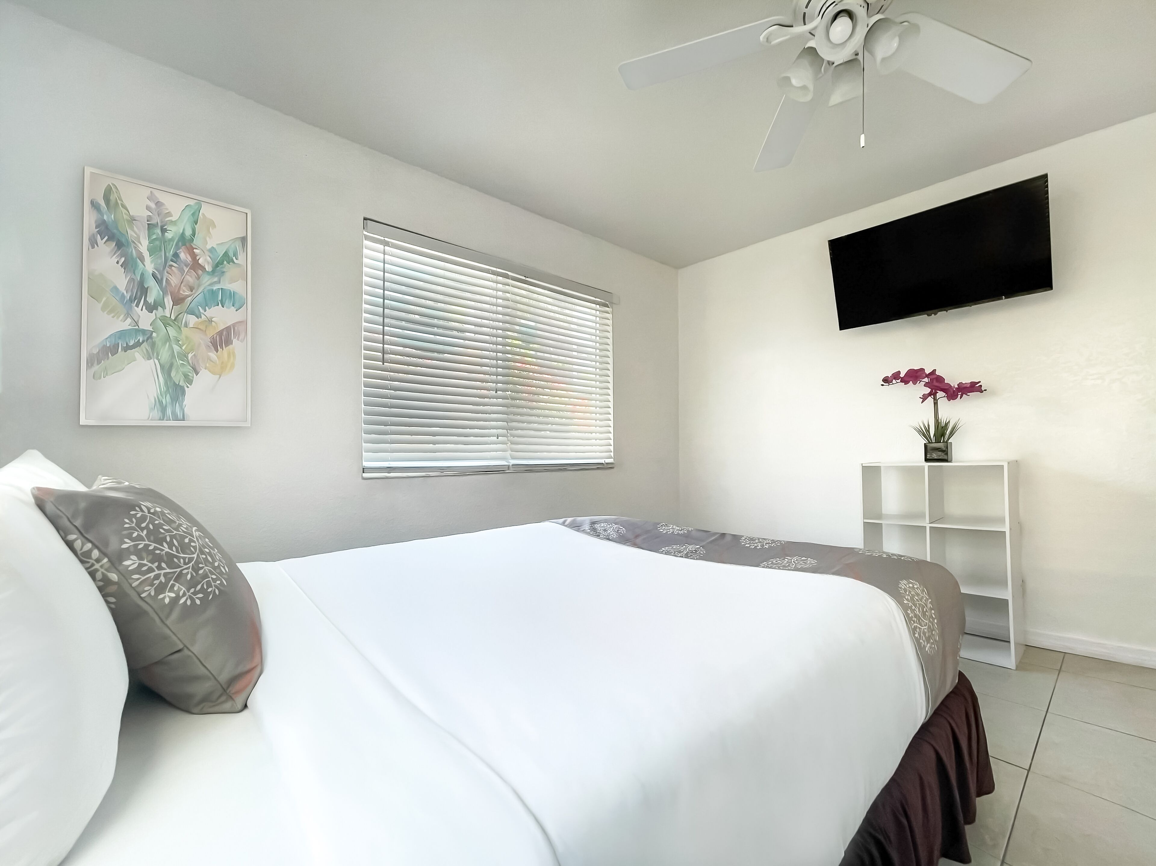 Een slaapkamer met een bed, een plafondventilator, een aan de muur bevestigde televisie en een raam met jaloezieën.