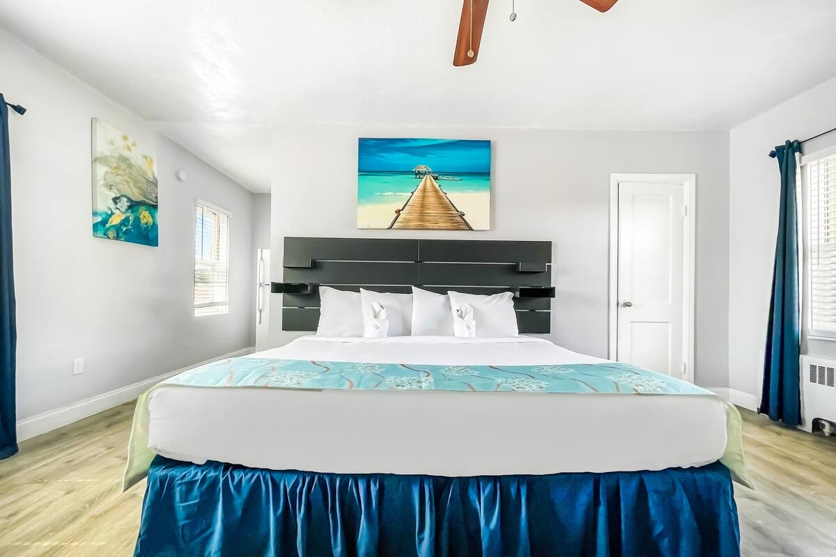 A hotel room with a large bed, a ceiling fan, a painting of a beach scene, and a window with curtains.