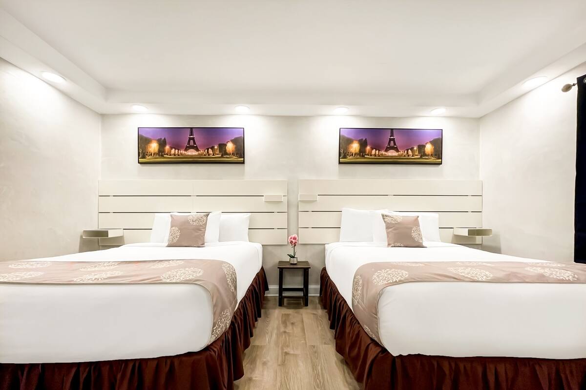 A hotel room with two beds, each with a headboard, a small table with a vase, and two wall-mounted paintings.
