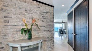 A modern hallway with a stone wall, a white console table with a vase of flowers, and a view to a living area with a sofa and a table. - Simply Spectacular 5th Floor Ocean View (Jacó)