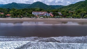 A beachfront hotel with multiple buildings, a sandy beach, and a mountainous backdrop. - Ground Floor Unit in Beautiful Beachfront Complex (Jacó)