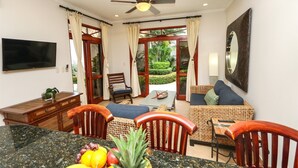 A room with a dining area, a sofa set, a TV mounted on the wall, and a view of a garden through the glass doors. - Charming Garden View Apt in Exclusive Community (Jacó)