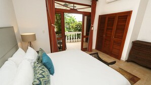 A bedroom with a large bed, a desk, and a chair. There is a window with curtains, a ceiling fan, and a wooden door. - Charming Garden View Apt in Exclusive Community (Jacó)