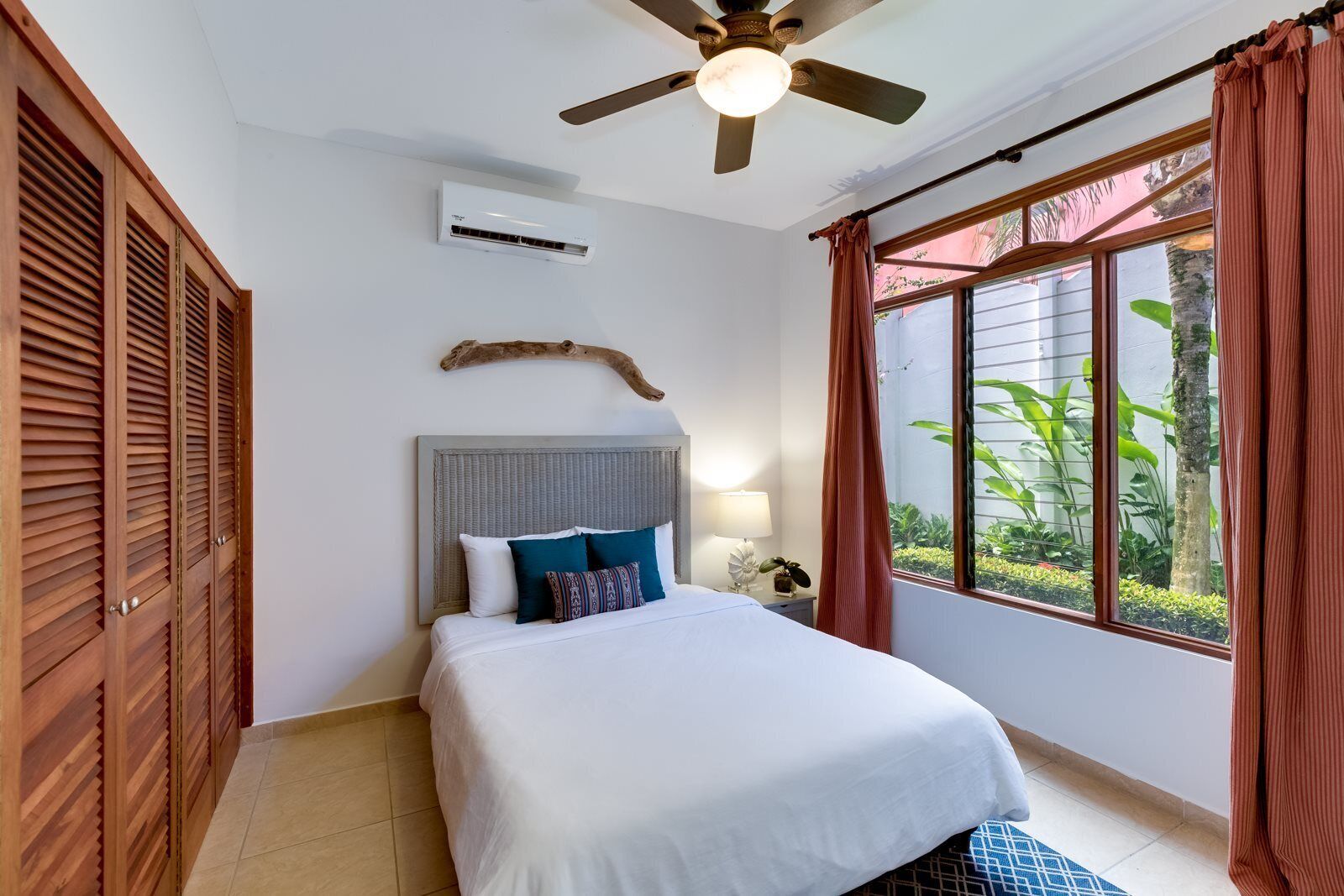 A bedroom with a bed, a ceiling fan, an air conditioning unit, a window with curtains, and a view of greenery outside.