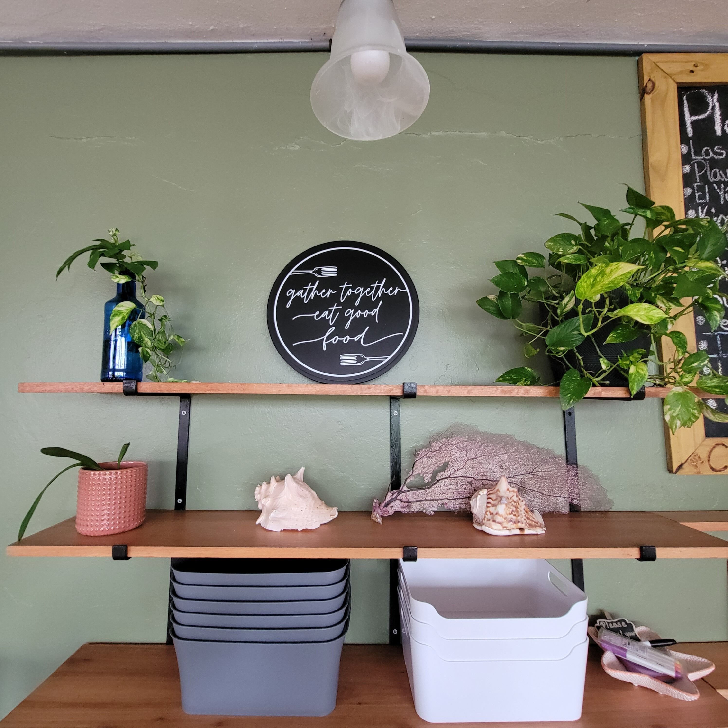 A kitchen counter with a chalkboard sign, a potted plant, a decorative shell, and storage bins.