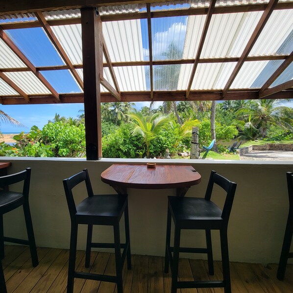 Un espacio de estar al aire libre cubierto con una mesa y sillas de madera, con vistas a un exuberante jardín y una playa.