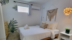 Un dormitorio con cama, una mesita de noche con una planta, aire acondicionado de pared y un colgador de macramé.