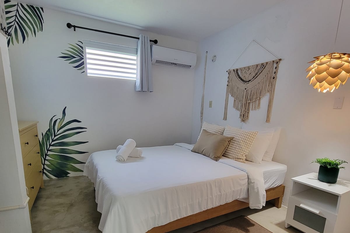 Un dormitorio con cama, una mesita de noche con una planta, aire acondicionado de pared y un colgador de macramé.
