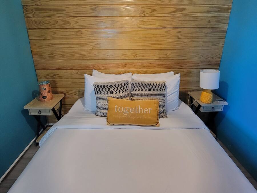 A neatly made bed with white linens, multiple patterned pillows, and a 'together' pillow. A wooden headboard and a hanging light fixture are visible.
