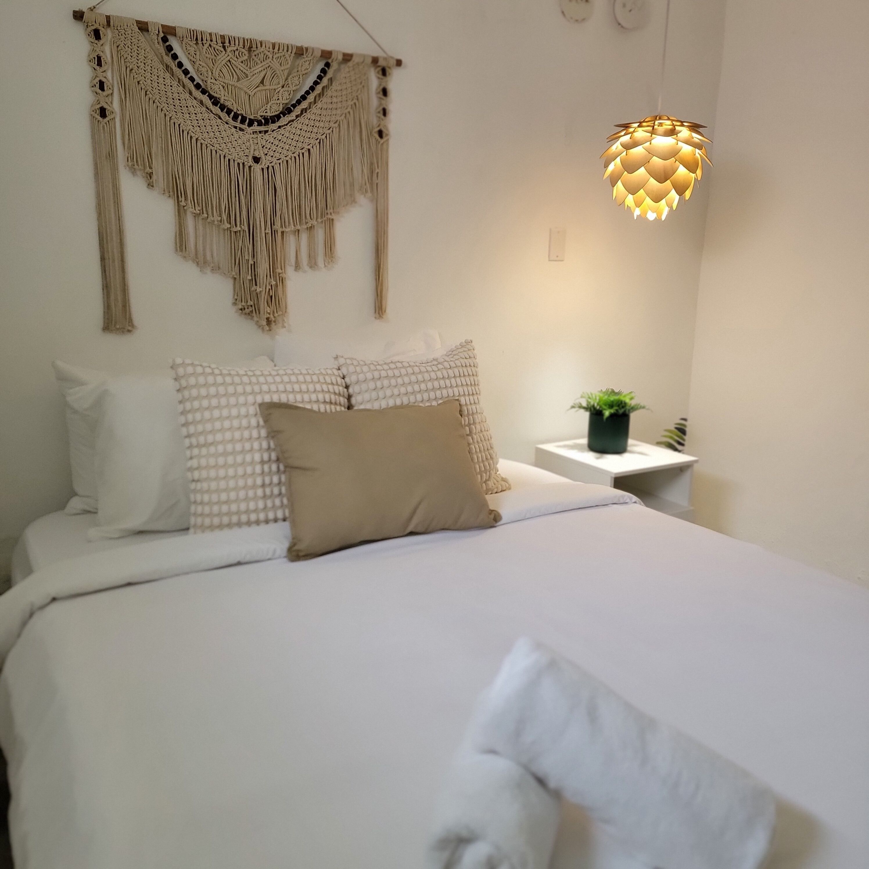 Une chambre à coucher avec un lit, des oreillers, une décoration murale en macramé, une plante sur la table de chevet et un luminaire suspendu.
