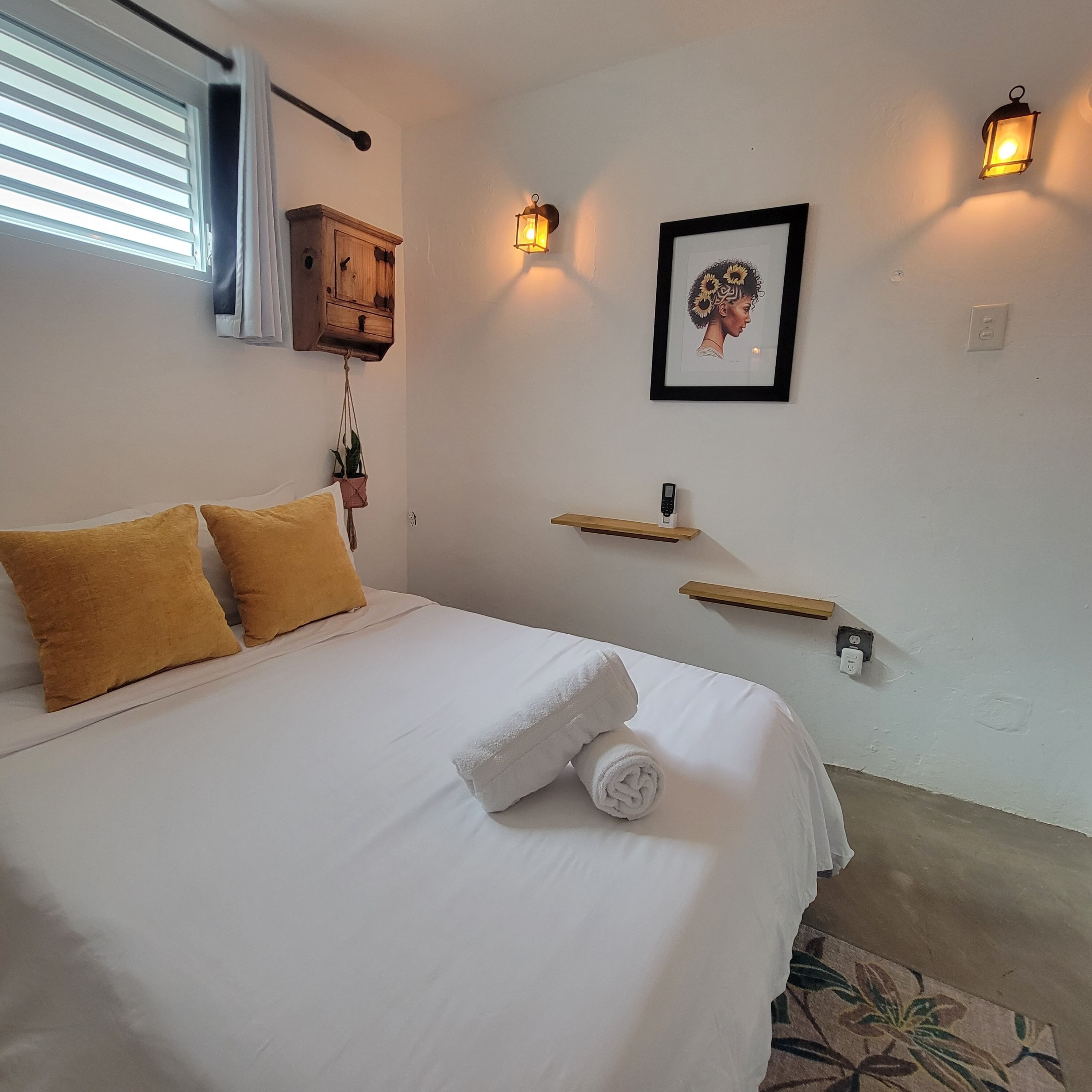 A neatly made bed with white and mustard pillows, a rolled white towel, a wooden cabinet, a framed artwork, and two wall-mounted lights.