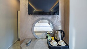 Habitació d'hotel amb un llit, una televisió penjada a la paret, un racó de cafè amb una tetera, tasses i aperitius, i un mirall rodó a la paret.