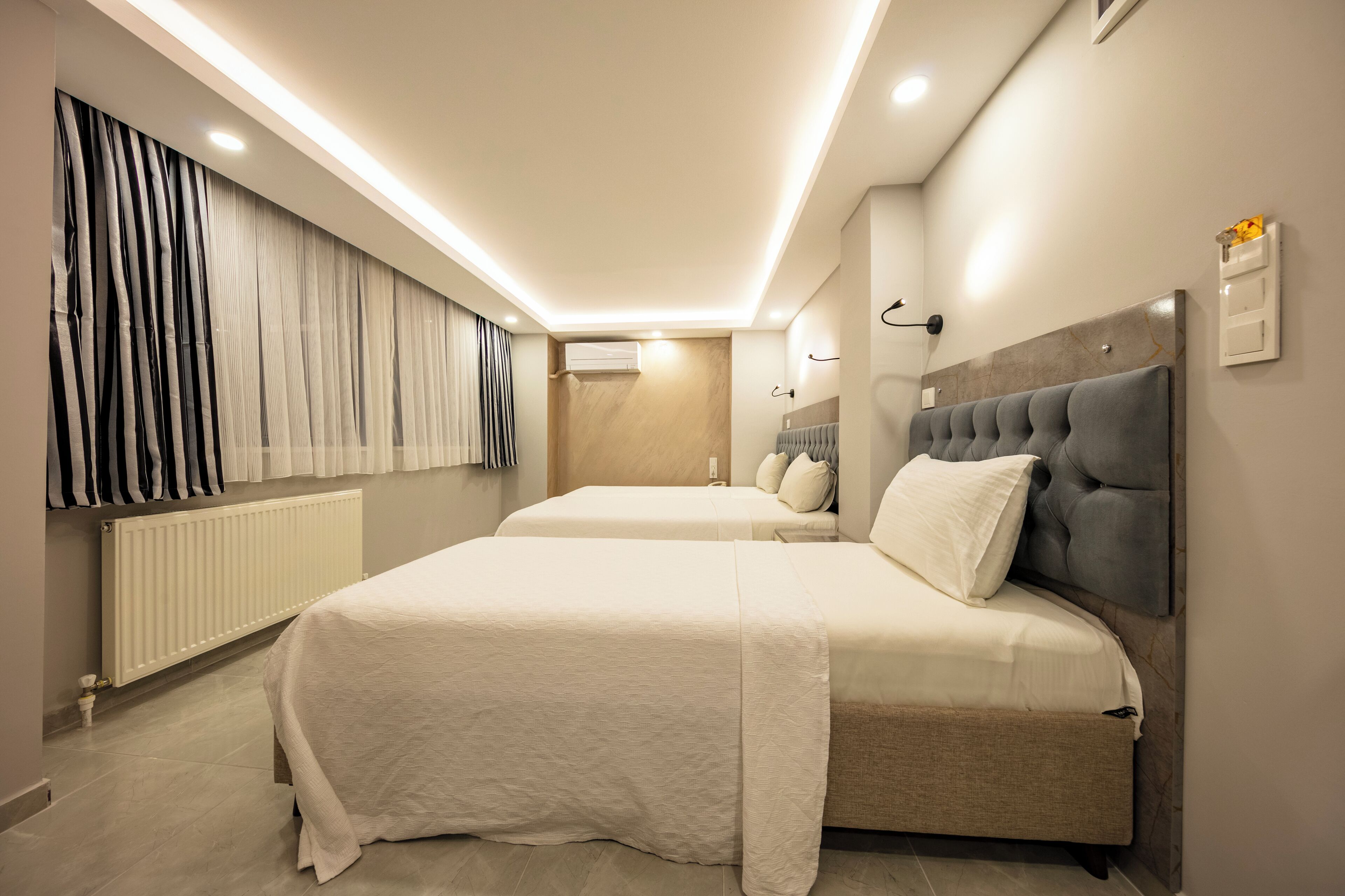 Una habitación de hotel moderna con dos camas, un ventanal con cortinas, un radiador y un aire acondicionado de pared.