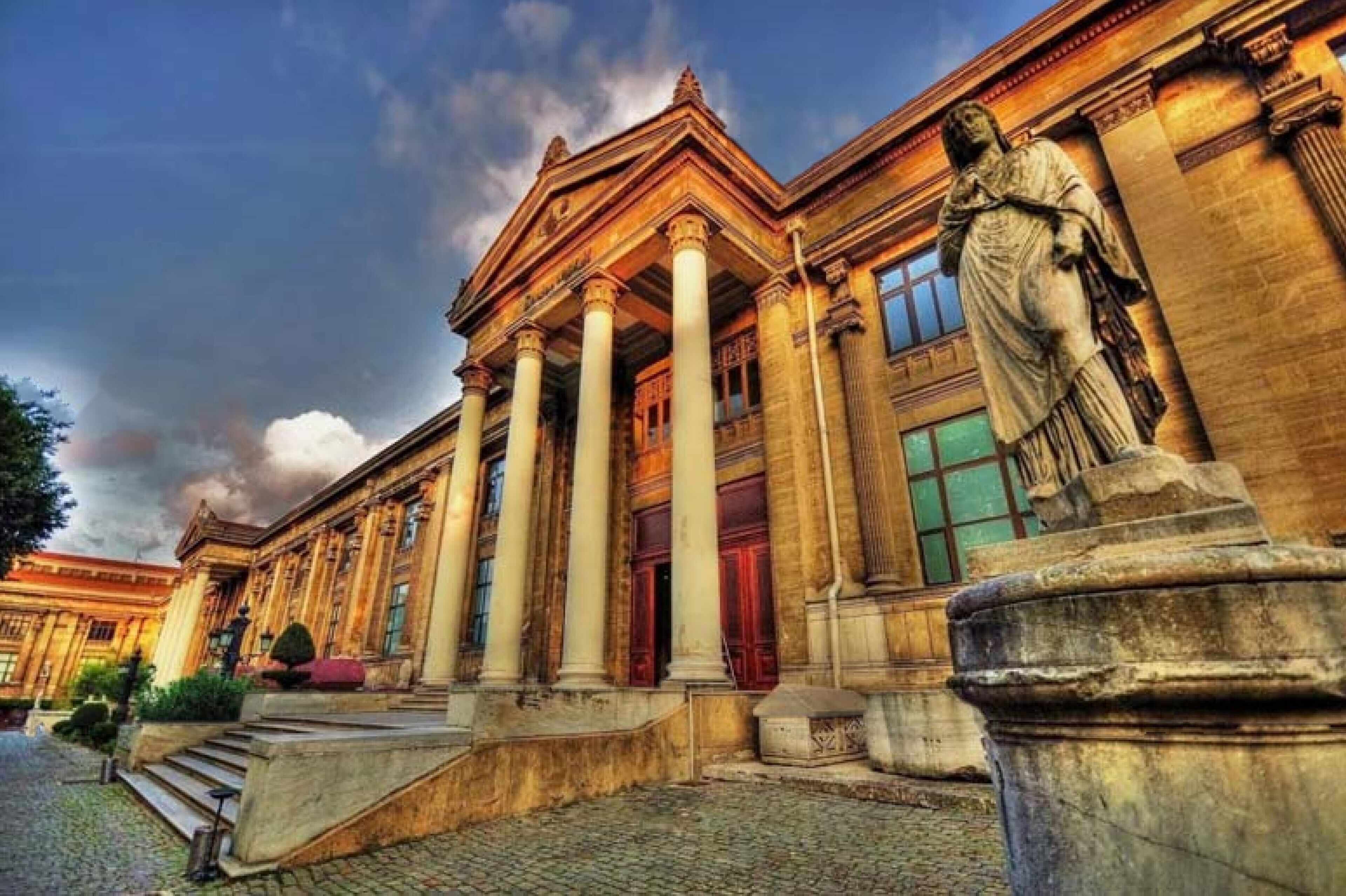 A classical building with a central entrance flanked by columns, a statue, and a cobblestone pathway.
