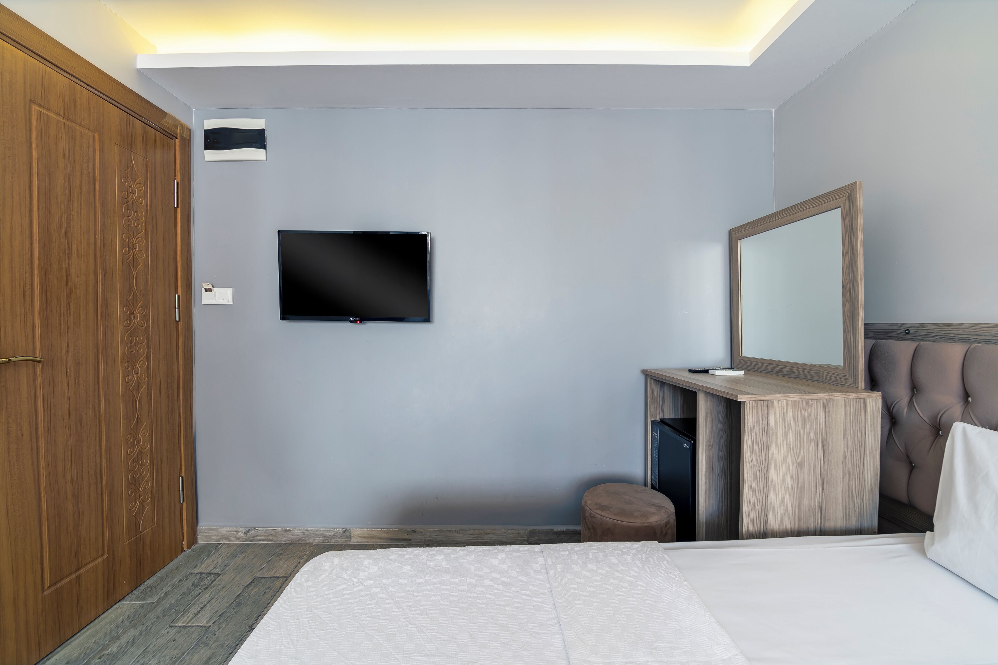 Una habitación de hotel moderna con una cama grande, un televisor de pantalla plana montado en la pared, un armario de madera, una tocador con espejo y un taburete pequeño.