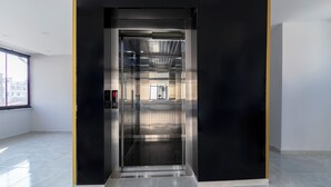 Un ascensor modern amb acabat metàl·lic i panell de vidre, ubicat en una estança àmplia i ben il·luminada amb un gran finestral.