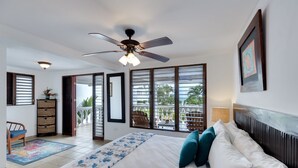 Une chambre à coucher comprenant un lit, un ventilateur de plafond, une fenêtre avec des stores, une chaise et une petite table.