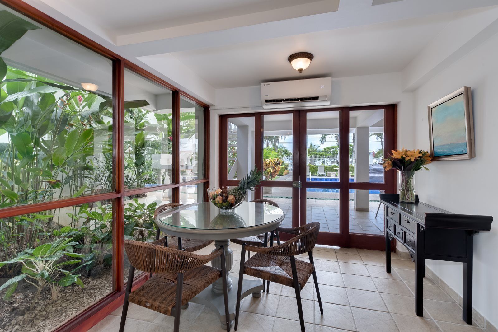 A room with a glass table, wicker chairs, a vase with flowers, an air conditioning unit, and a view of a pool and greenery outside.