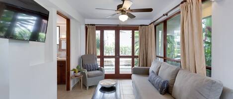 Amplia sala de estar con ventilador de techo, televisor de pantalla plana, sofá, sillón y una mesita con planta.