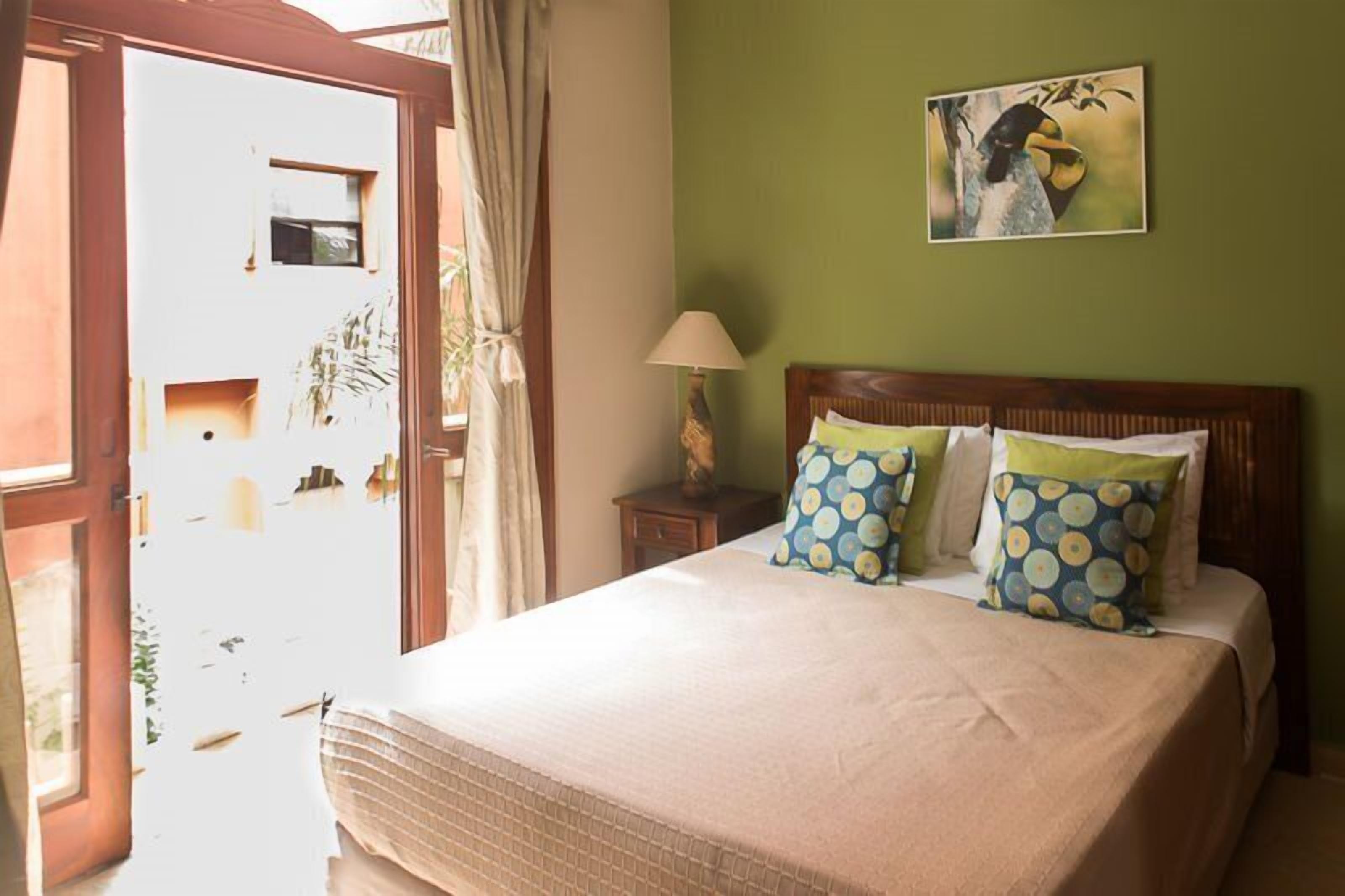 Quarto com cama, mesa de cabeceira, luminária, quadro e janela com cortinas.