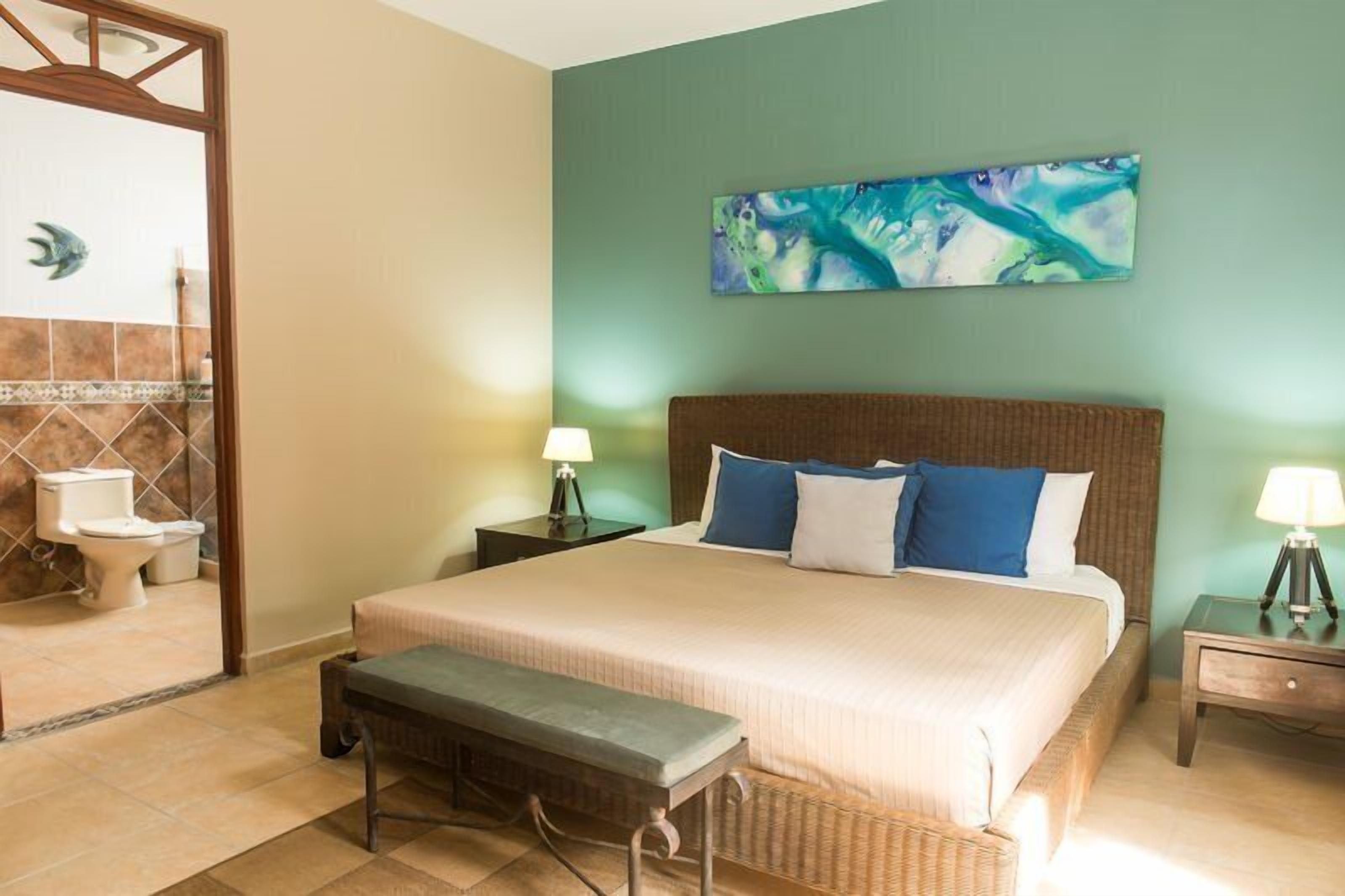 A hotel room with a large bed, a bench, two bedside tables with lamps, a bathroom with a toilet and sink, and a green abstract painting on the wall.