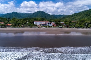 A beachfront hotel with multiple buildings, a sandy beach, and a mountainous backdrop. - Lpb-3f (Jacó)