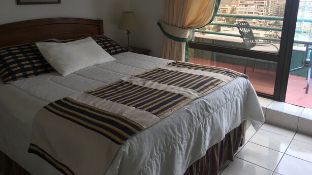 A hotel room with a bed, a nightstand, a lamp, a window with curtains, and a balcony with a chair. - Apart Hotel Dos Providencia (Santiago)