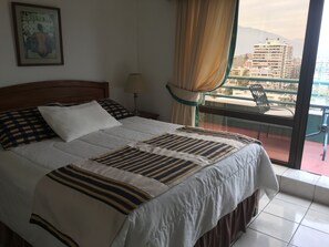 A hotel room with a bed, a nightstand, a lamp, a window with curtains, and a balcony with a chair. - Apart Hotel Dos Providencia (Santiago)