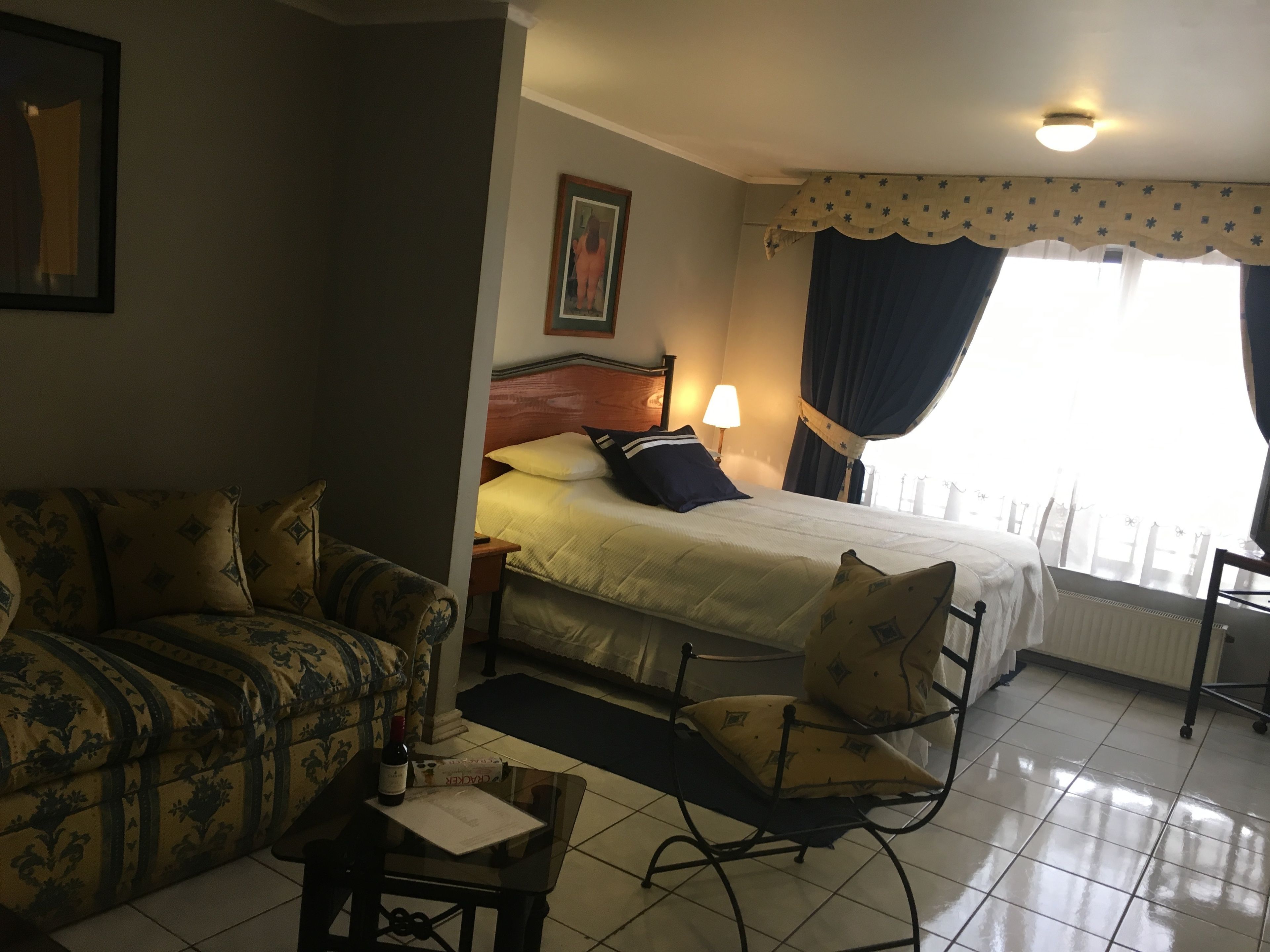 Habitación de hotel con cama, sofá, sillón, mesa con una botella y un menú, y ventana con cortinas.