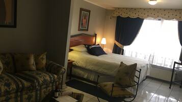 Habitación de hotel con cama, sofá, sillón, mesa con una botella y un menú, y ventana con cortinas.