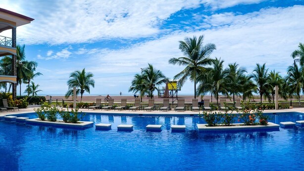 A swimming pool with lounge chairs and palm trees, surrounded by hotel buildings. - Terrific 2BR 1st Floor Unit is Sure to Please (Jacó)