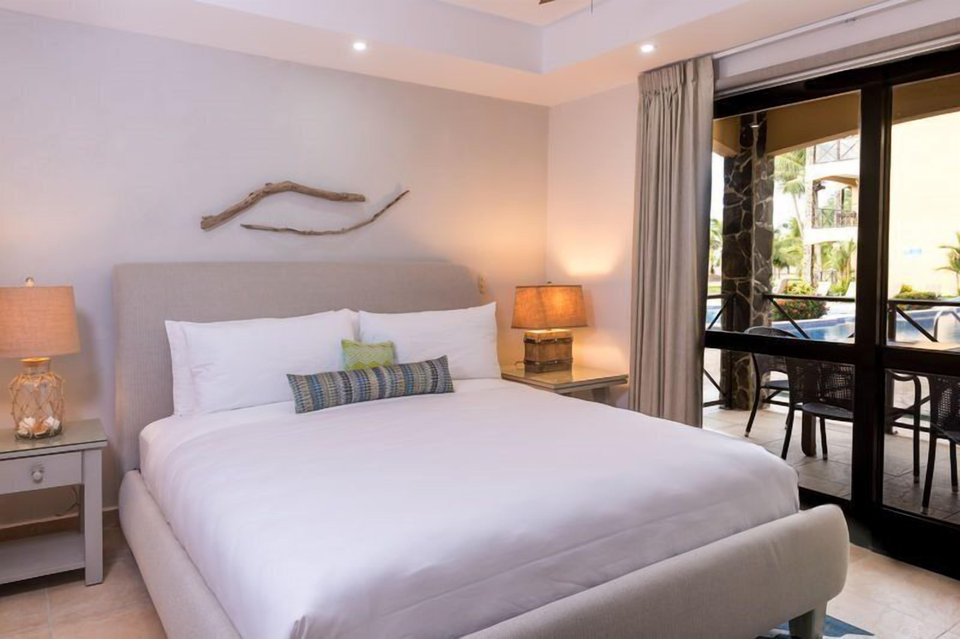 A hotel room with a large bed, two bedside tables with lamps, a view of the outdoor area through a glass door, and a decorative branch on the wall.