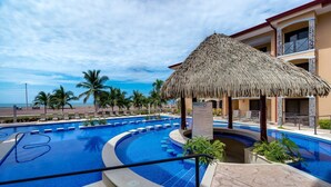 A pool area with a thatched roof structure, palm trees, and a view of the beach. - Oceanfront Penthouse / Upscale Roof Terrace (Jacó)