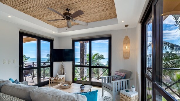 A spacious living room with a large sofa, a coffee table, and a chair. There are large windows offering a view of the ocean. A ceiling fan is visible, and the room has a modern design with a woven ceiling. - Oceanfront Penthouse / Upscale Roof Terrace (Jacó)