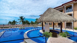 A pool area with a thatched roof structure, palm trees, and a view of the beach. - Amazing Seaside Penthouse With Rooftop Terrace (Jacó)