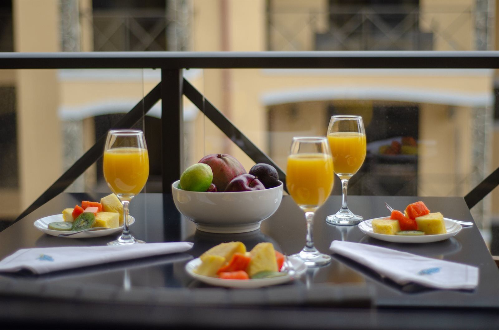 A breakfast setting with a bowl of fruit, two glasses of orange juice, and plates of fruit on a table with a view of buildings.