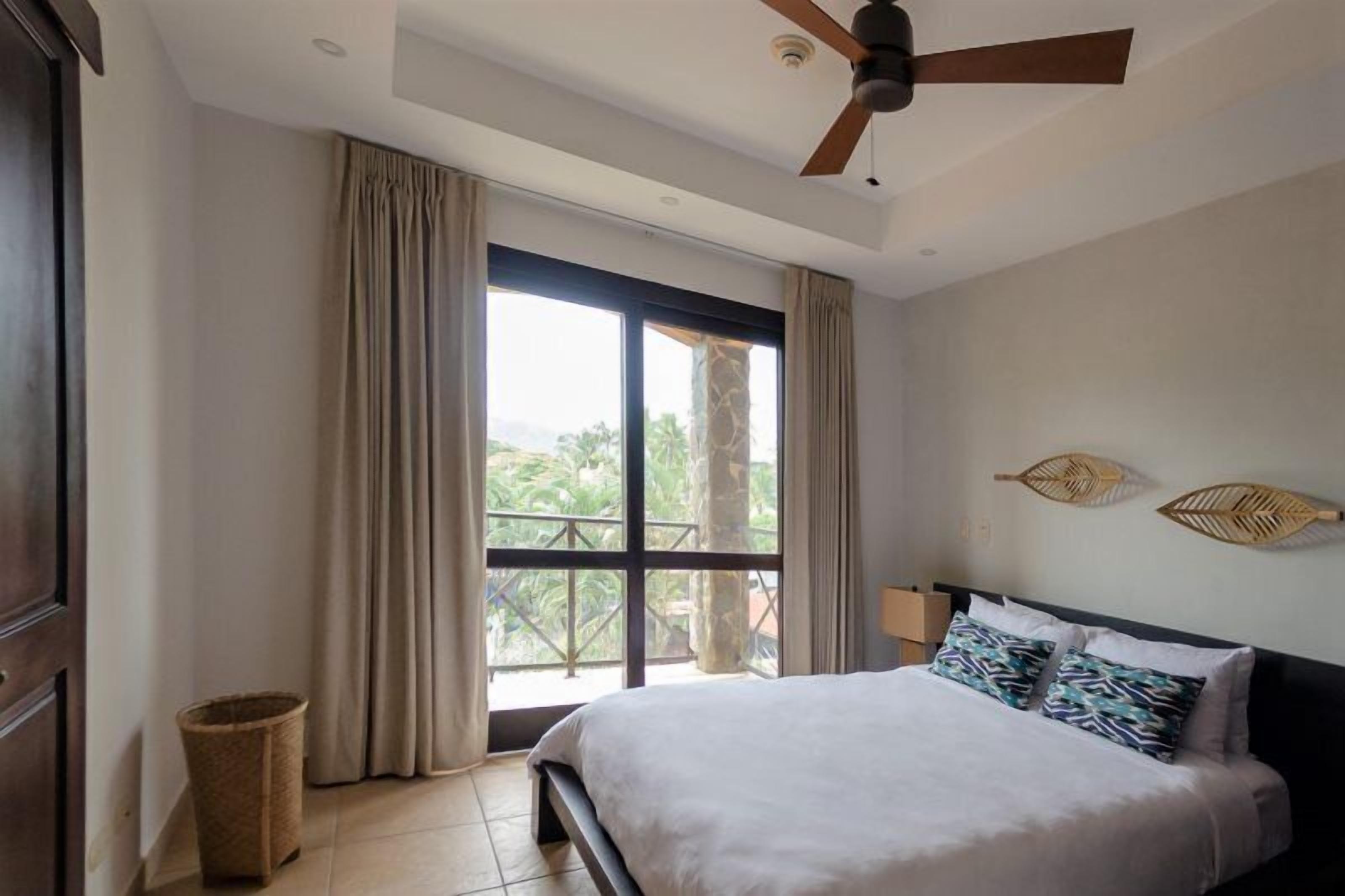 A bedroom with a bed, a ceiling fan, a window with curtains, and a view of greenery outside.